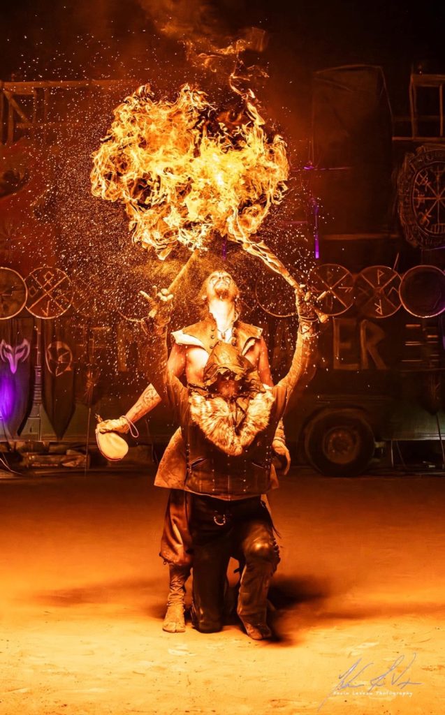 A hooded fire priestess raises two blazing pentagram wheels
